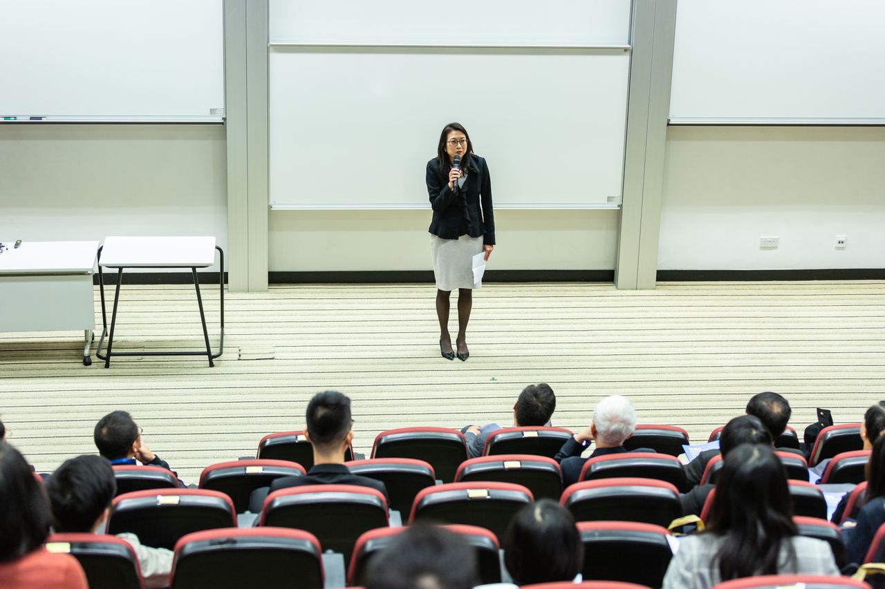 Offerings Female speaker presenting to an audience in a modern auditorium setting.